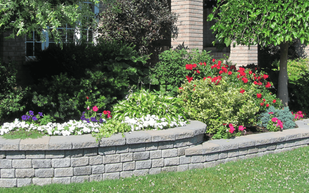 A garden showcasing a stone flower bed with vibrant flowers and a manicured tree in front of a brick house.